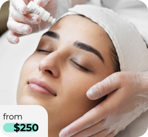 Close-up of a woman receiving a facial treatment; a skincare professional wearing gloves applies product from a tube to her forehead while she rests with eyes closed, wearing a head wrap