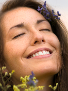 Close-up of a smiling woman with eyes closed, freckles, and purple flowers tucked into her hair, with yellow wildflowers in the foreground and a soft sky in the background.