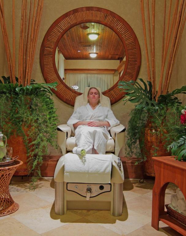 A woman in a white spa robe sits with her eyes closed in a cream-colored pedicure chair, a towel on her lap, in a tranquil spa room surrounded by a large round wooden mirror and lush green plants.