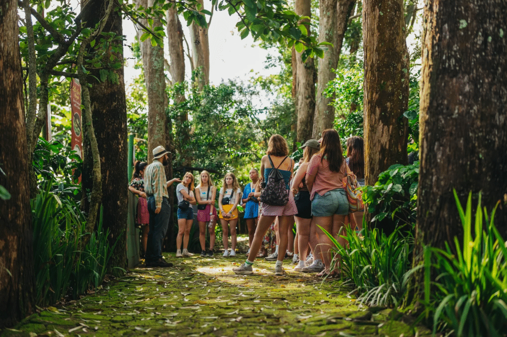 A tour guide wearing a hat speaks to a group of casually dressed young people with backpacks on a mossy forest path, surrounded by tall trees and lush green foliage.