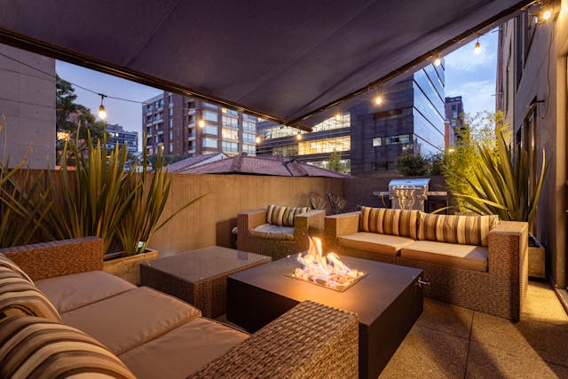 Rooftop urban lounge with beige wicker sofas arranged around a square fire pit, string lights overhead, tall planters with greenery, and a city skyline in the background at dusk.